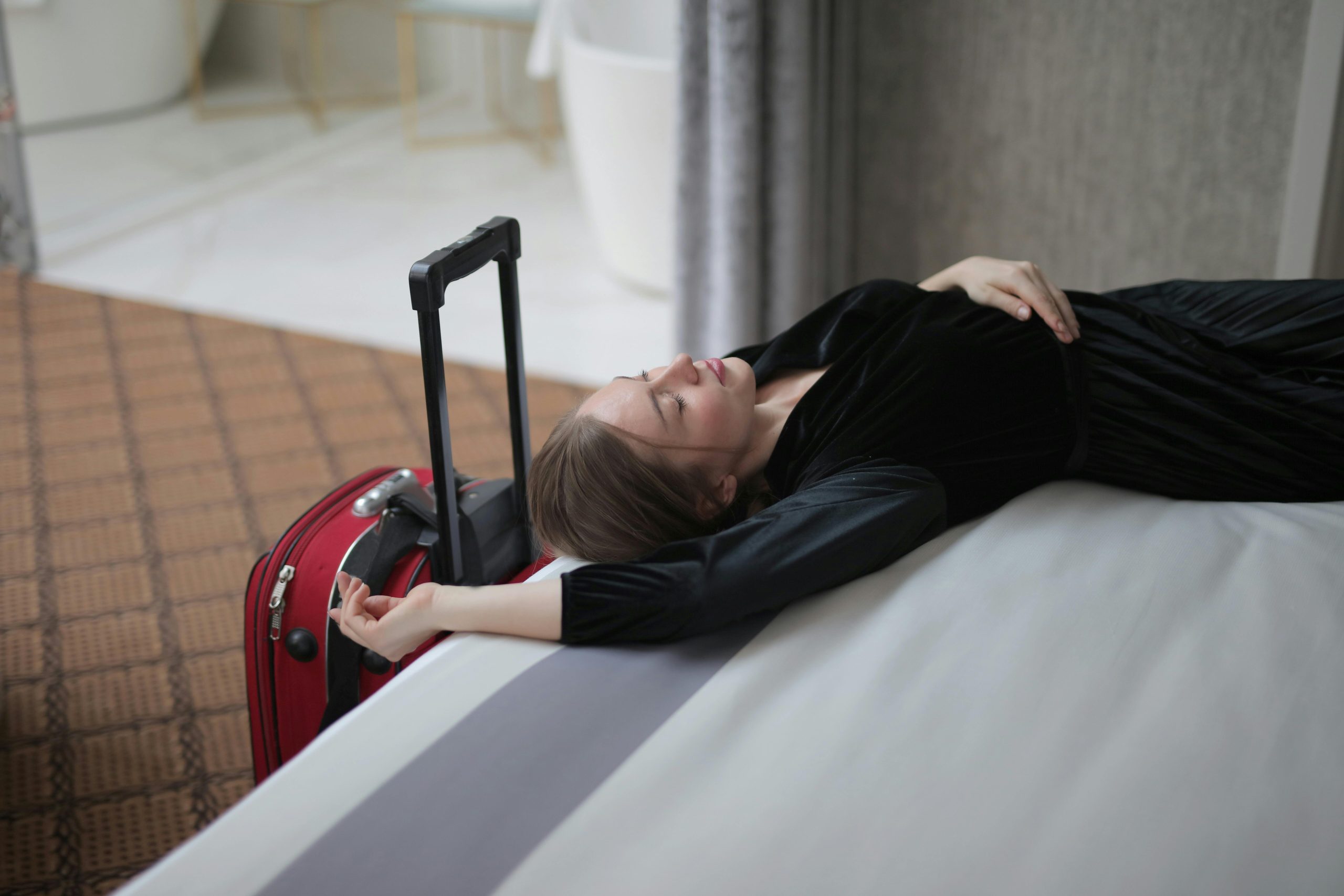 Woman lying on a hotel bed next to a red rolling suitcase after a long flight