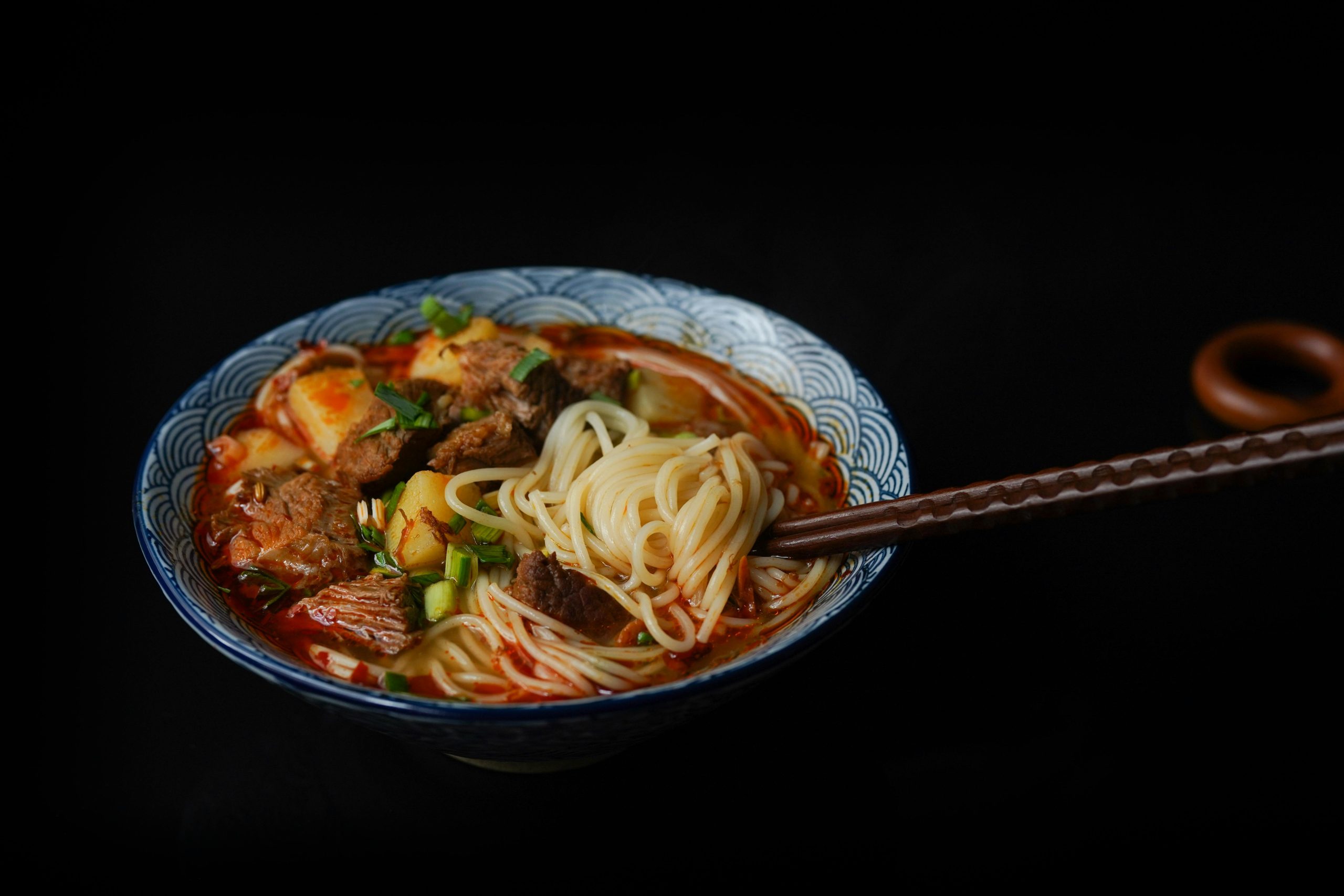 A bowl of spicy noodle soup with beef and scallions in a traditional blue-pattern Chinese bowl