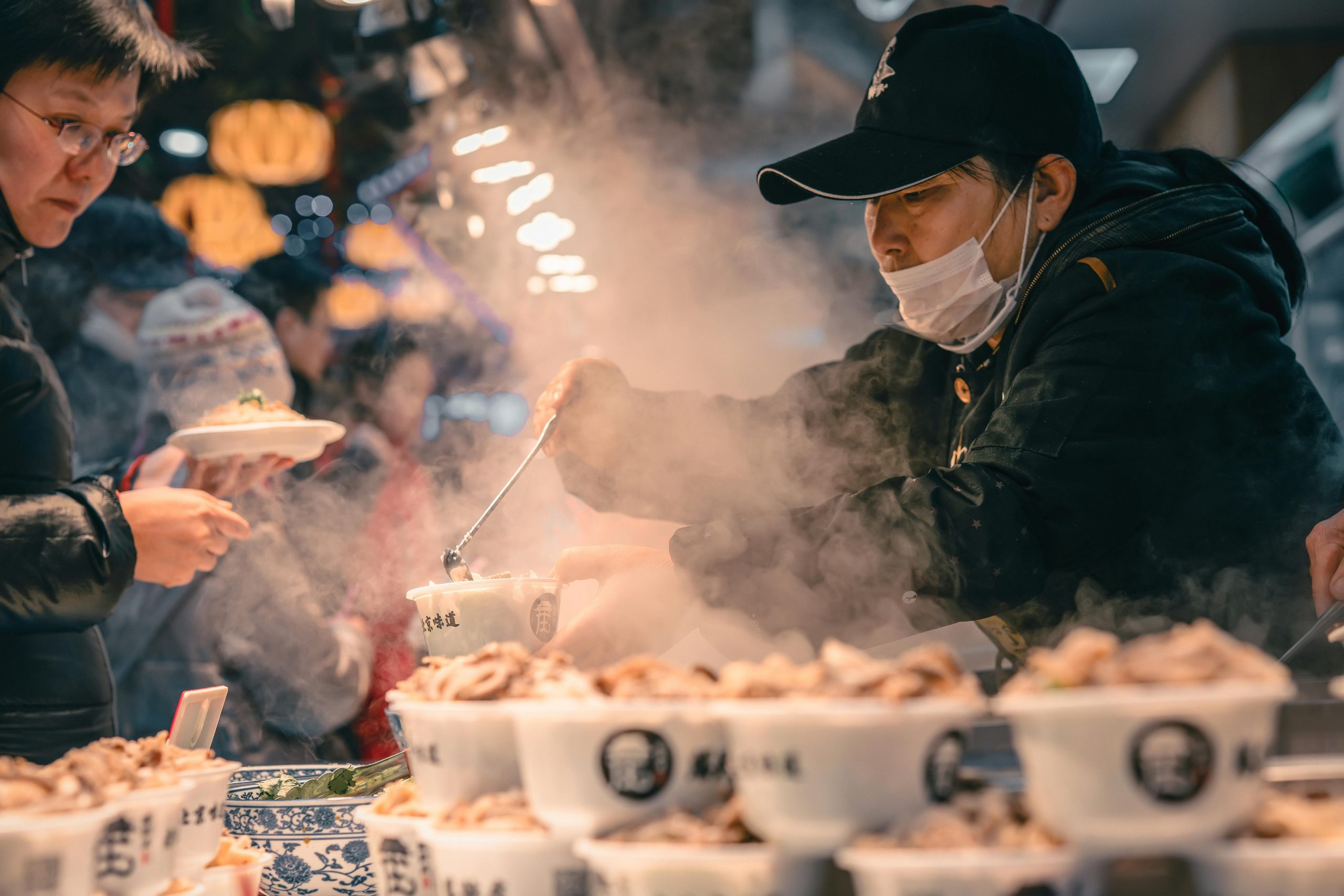 A vendor serves steaming bowls of food at a busy Chinese street stall while a customer reaches for a dish