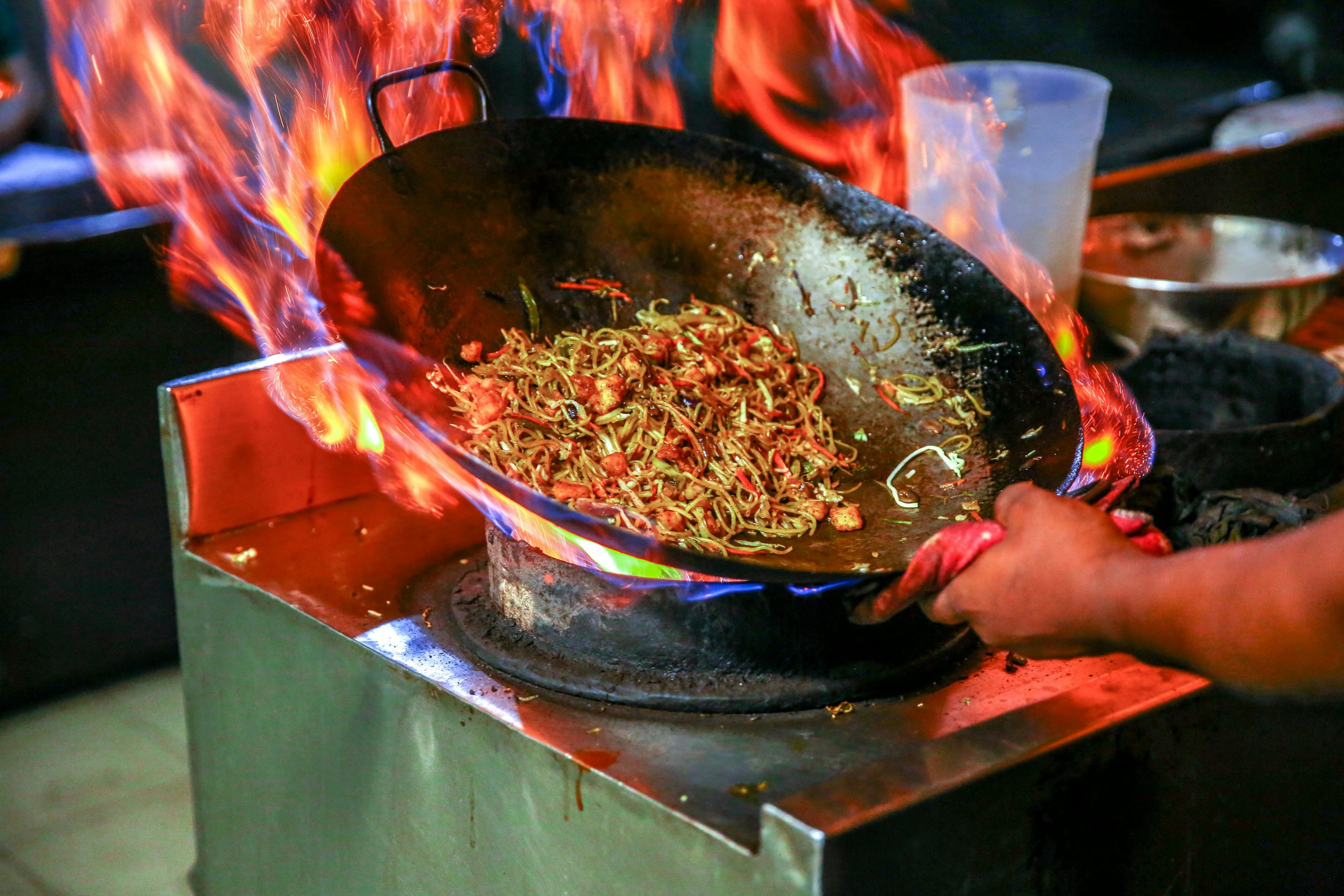 Noodles being tossed in a wok with dramatic flames shooting up around the pan