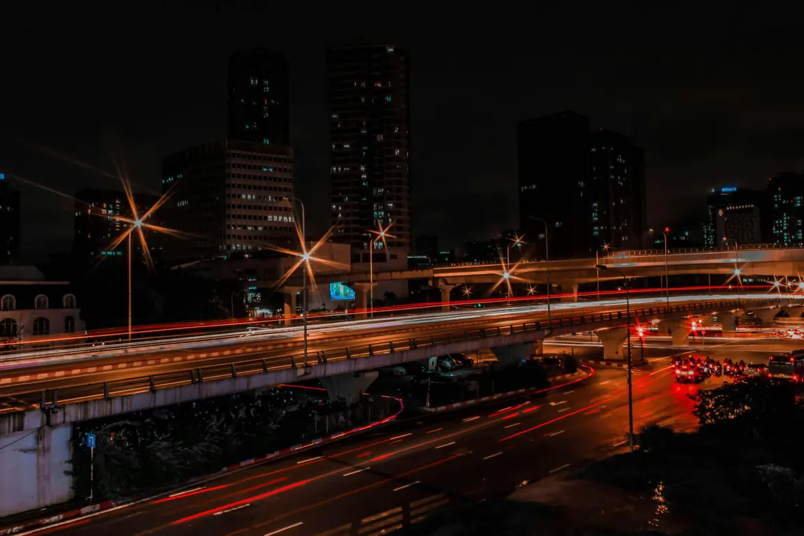 Night highway cutting through an Asian city skyline with car light trails and illuminated high-rises